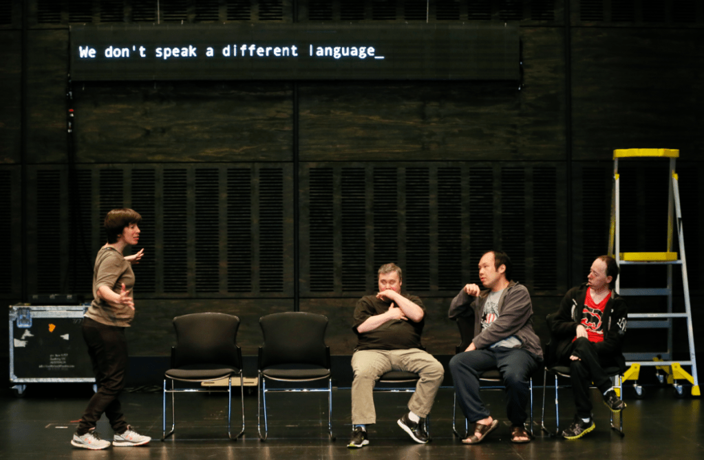 A production photo from The Shadow Whose Prey the Hunter Becomes. A woman stands on the left gesturing to a group of three men sitting in chairs on the right. Above them is a screen that reads "We don't speak a different language."