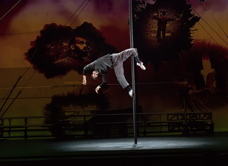 The 7 Fingers performer hanging sideways on a pole