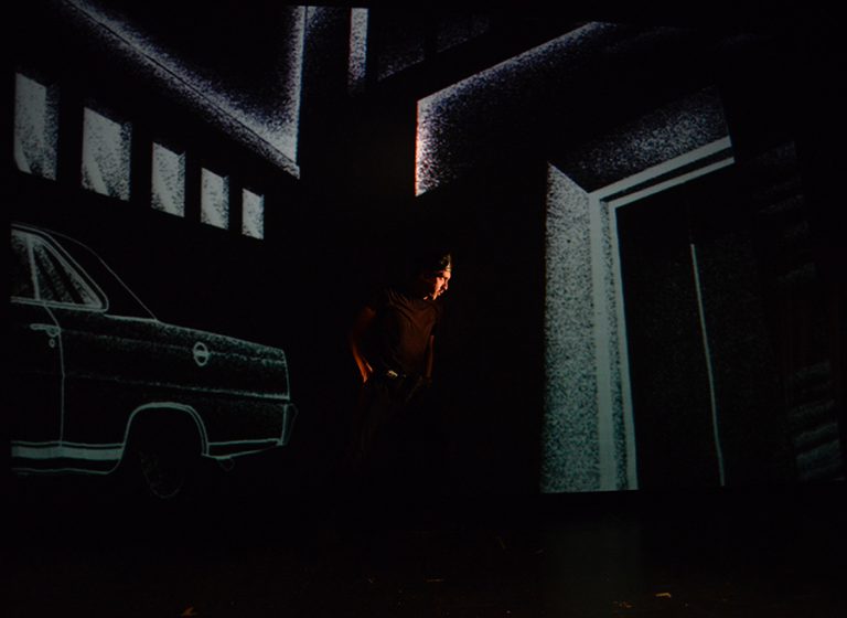 Man standing in middle of dark scene with projections of a sketched scenery of a car and buildings behind him in Plata Quemada