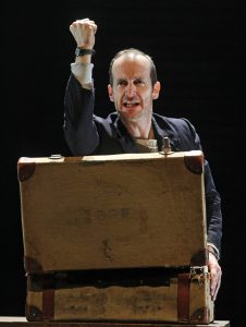 Dennis O'Hare performing in An Illiad. Man leaning over on briefcase with fist in the air.