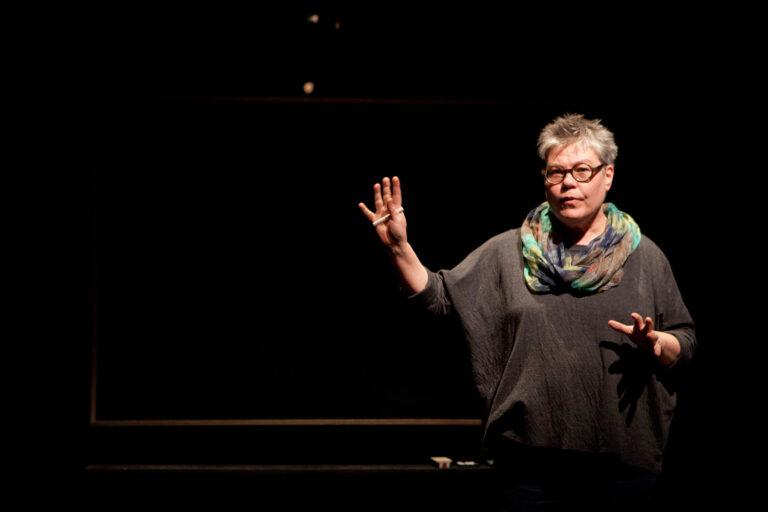 Alanna Mitchell performing a monologue on a dark stage in front of a blackboardin Sea Sick