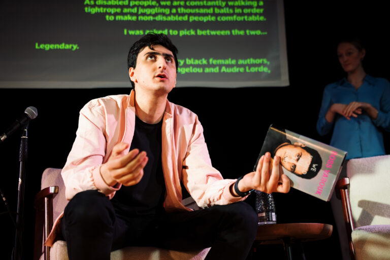 Actor AArian sitting in a chair, holding a book speaking with arms open.
