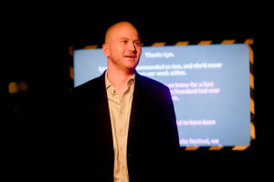 Samuel Brewer Standing in a suit and shirt with white collar speaking i front of a screen.