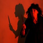 One actor holding a knife in front of a red screen, shadows projected onto the screen